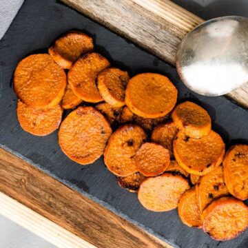 Roasted sweet potato slices are elegantly arranged on a black slate serving board, ideal for Thanksgiving sides. A silver spoon rests nearby, ready to serve and impress your in-laws. The setup is completed with a wooden tray and a touch of gray napkin peeking through.