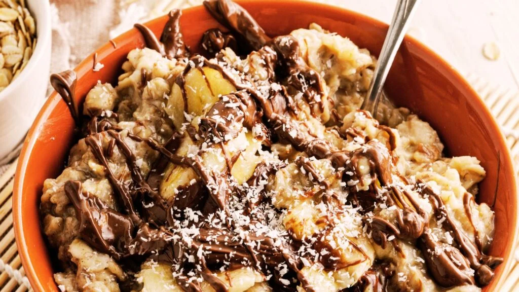 A bowl of oatmeal topped with banana slices, chocolate drizzle, and shredded coconut is shown. A spoon rests inside the bowl, as if drafting the perfect bite, and the dish is set on a wooden table. A small bowl of seeds is partially visible in the background.