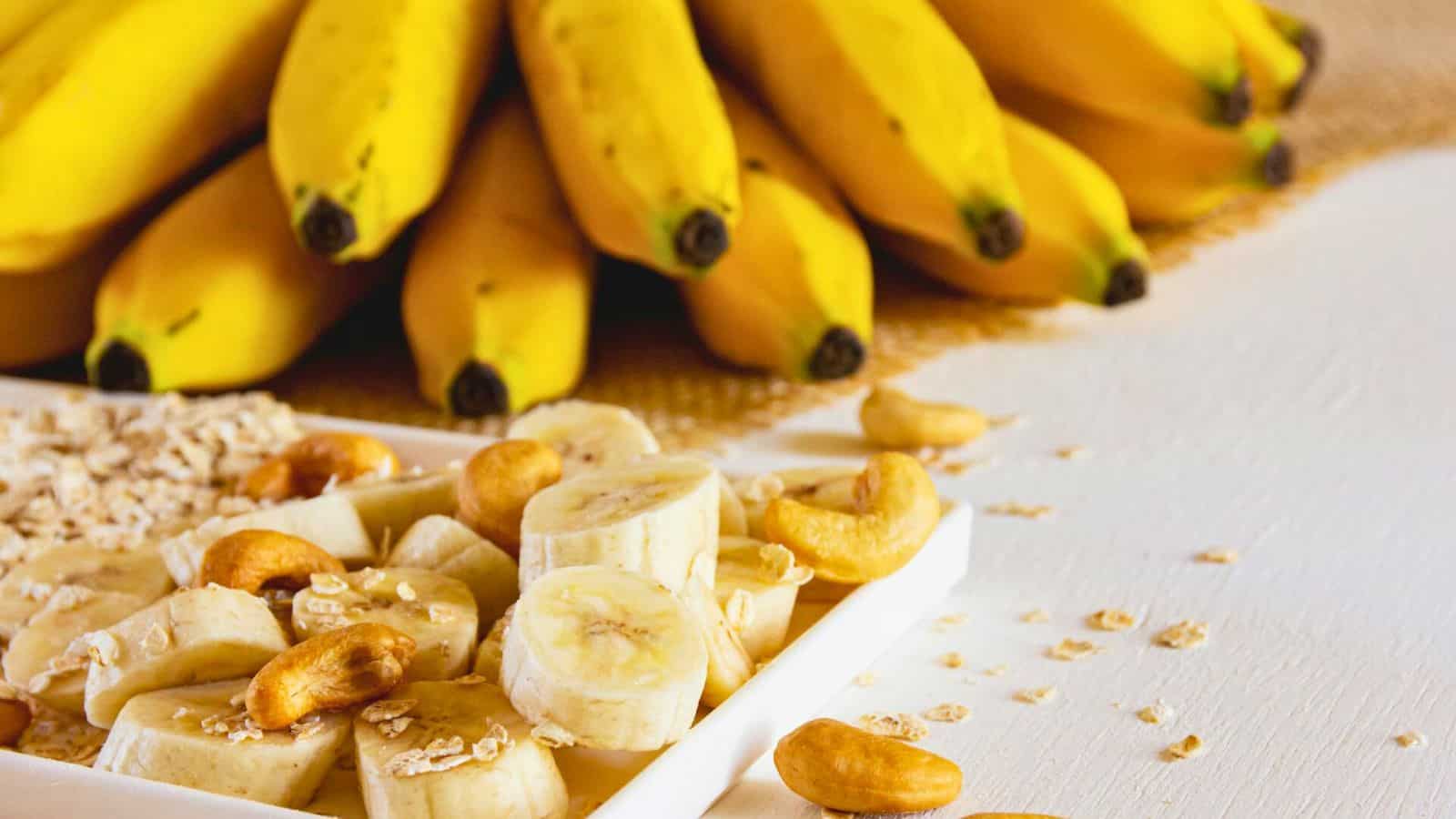 A white plate with sliced bananas, cashews, and oats sits on a white surface. In the background, there is a bunch of ripe bananas on a burlap cloth. Some oats and cashews are scattered around the plate.