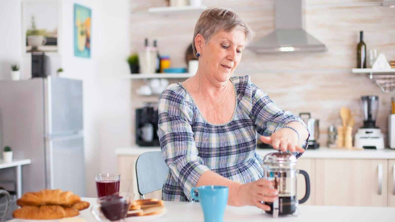 A woman with short gray hair is pressing down on a French press coffee maker in a kitchen. She is seated at a table with croissants, toast, and a blue mug. The kitchen is modern with shelves and various items in the background.