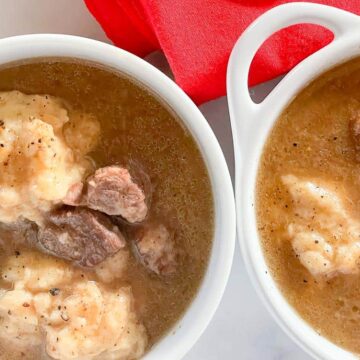 Two bowls of beef stew with chunks of meat and dumplings are placed side by side. The stew has a rich, brown broth and is lightly seasoned. A red cloth is partially visible in the background.