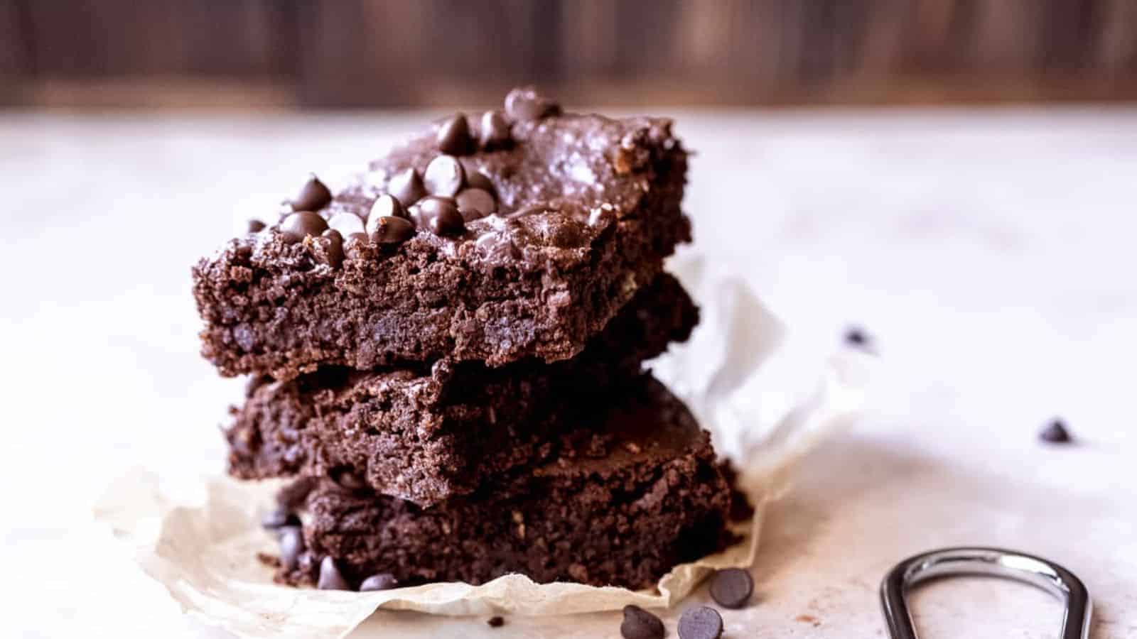 A stack of three chocolate brownies with chocolate chips on top rests on a piece of parchment paper. Some chocolate chips are scattered around. The background is softly blurred.