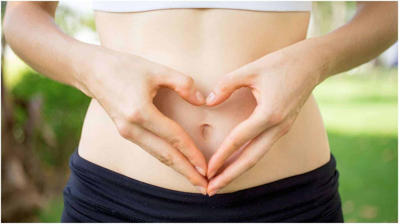 A person wearing a white crop top and black pants forms a heart shape with their hands over their bare midriff. The background is blurred and green, indicating an outdoor setting.