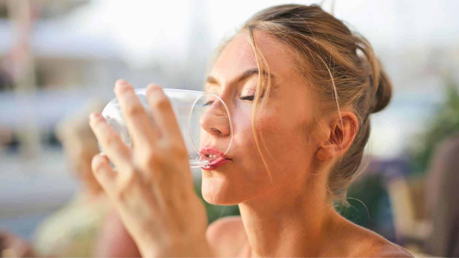 A woman with blond hair in a casual updo is sitting outside and drinking water from a clear glass. She has her eyes closed and appears to be enjoying the refreshment. The background is out of focus, suggesting an outdoor setting.