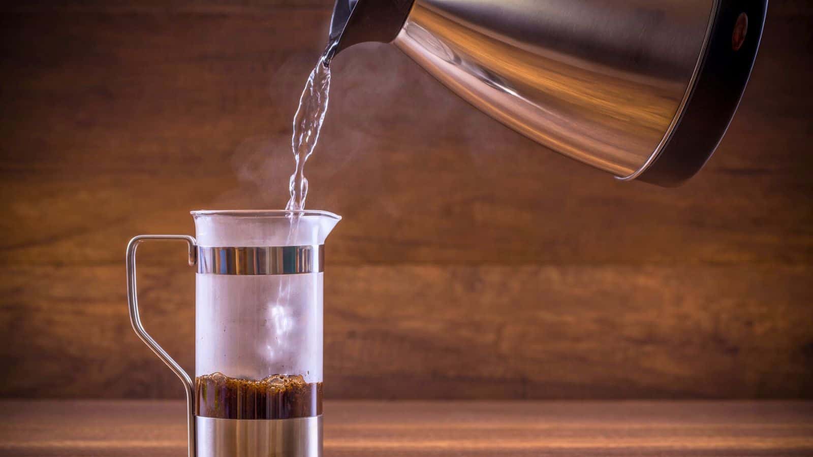 Hot water is being poured from a metal kettle into a French press containing coffee grounds on a wooden surface. Steam rises from the kettle.