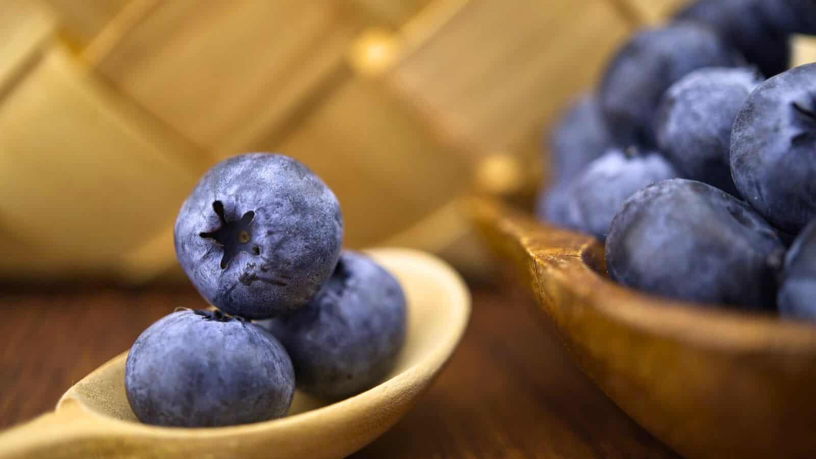 Close-up of fresh blueberries on a wooden spoon in the foreground, with a basket weave pattern blurred in the background. More blueberries are partially visible in a wooden bowl to the right.