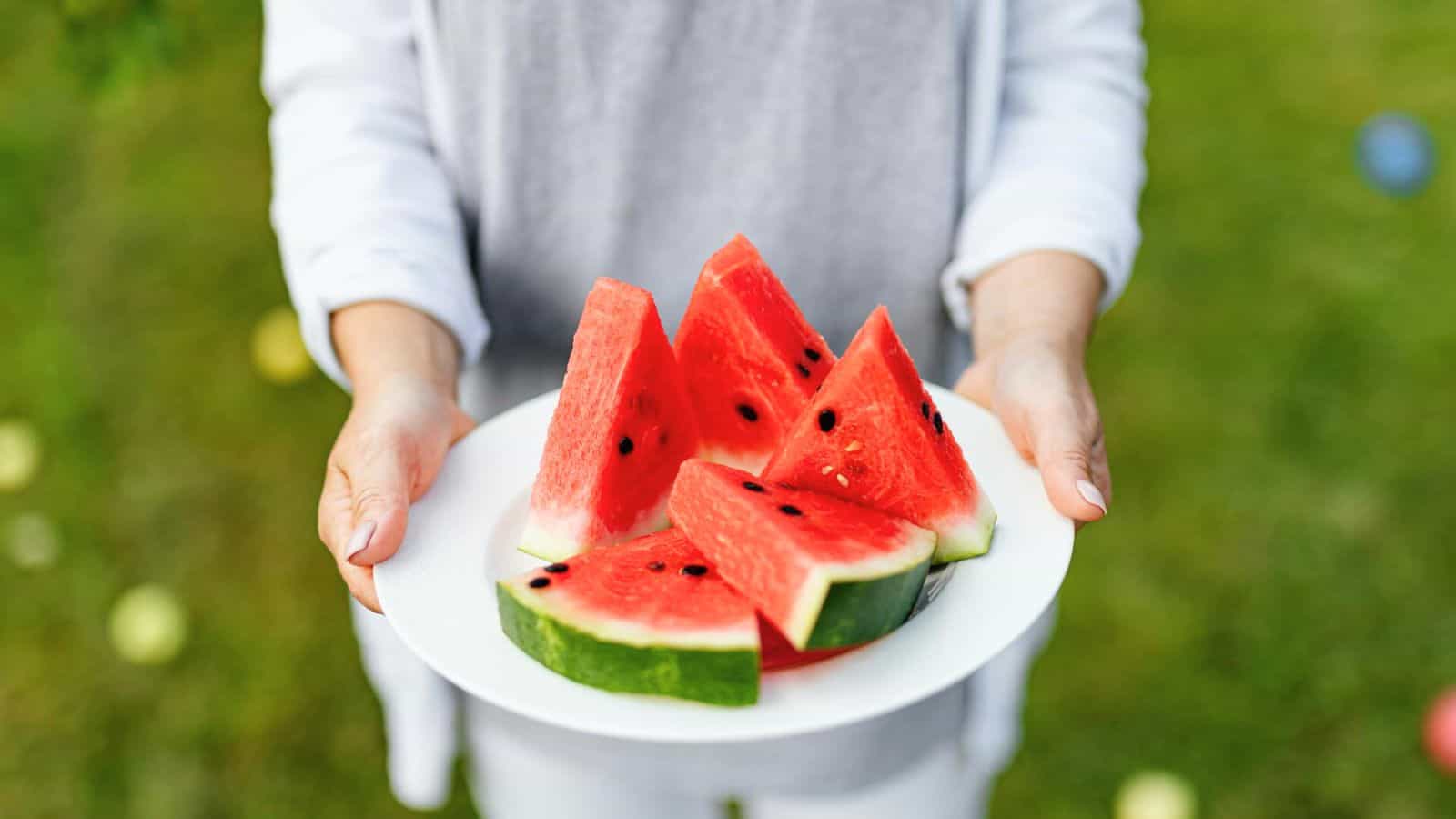 A person wearing a white shirt holds a white plate with sliced watermelon. The watermelon is red with black seeds and green rind. The background is a grassy area with some blurred round objects.
