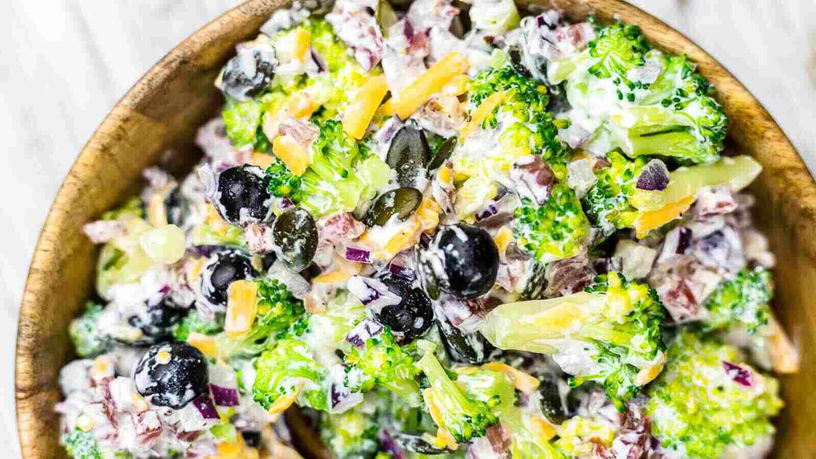 A bowl of broccoli salad with florets, sliced red onions, shredded cheddar, black olives, and creamy dressing garnished with sunflower seeds makes a delightful addition to any Thanksgiving dishes spread.