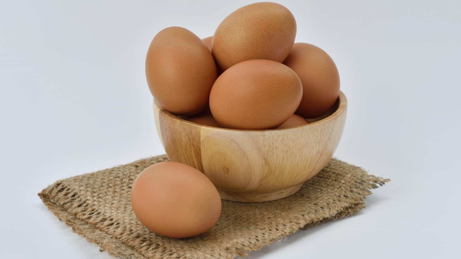 A wooden bowl filled with several brown eggs sits on a piece of burlap fabric, with one egg resting outside the bowl. The background is plain white.