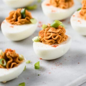 A close-up of deviled eggs on a white surface showcases a keto-friendly delight. Each half egg boasts a creamy filling topped with vibrant red paprika and tiny green onion pieces, beautifully arranged with chopped green onions sprinkled around for an elegant finish.