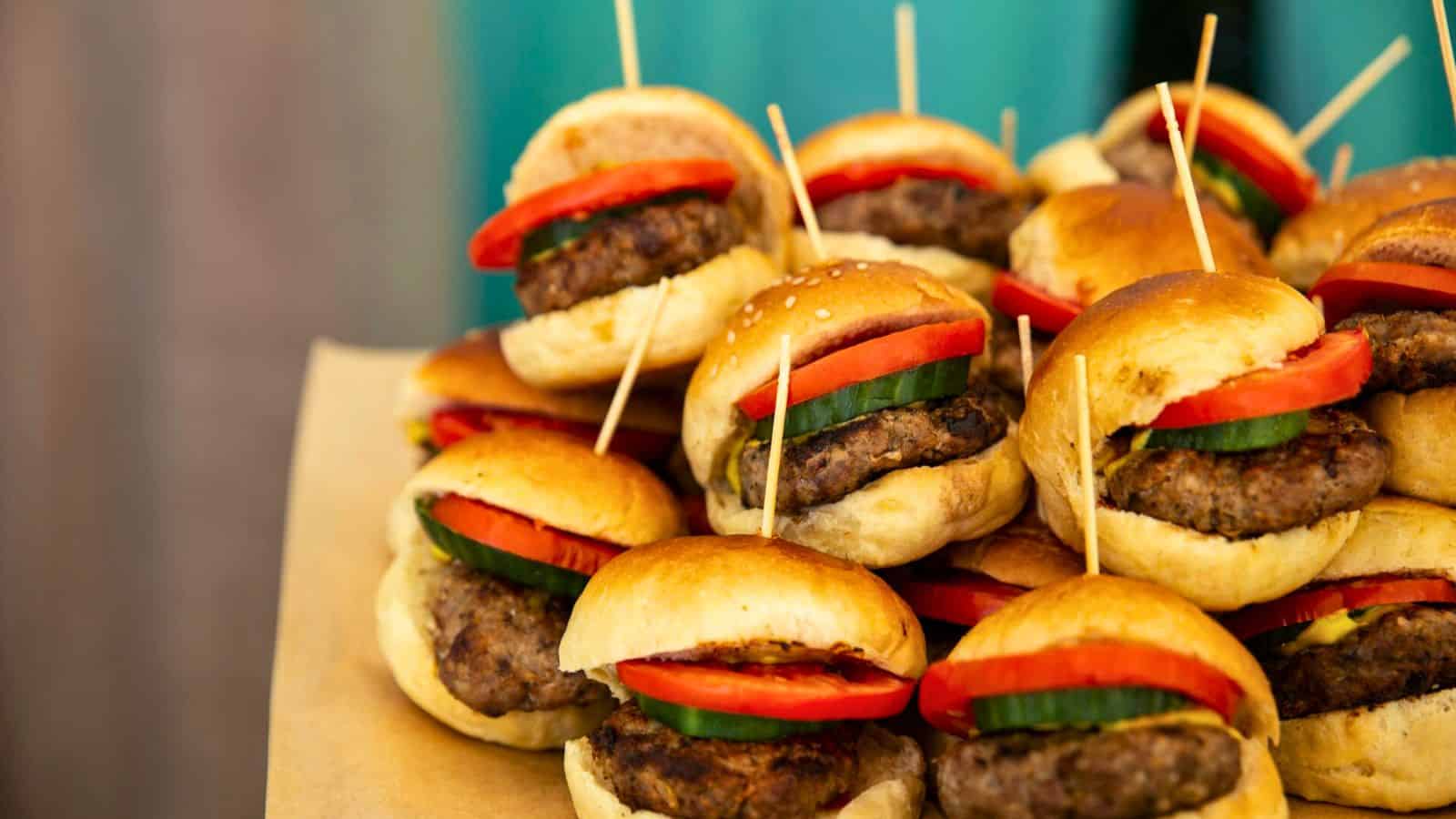 A stack of mini burgers on a tray, featuring juicy beef patties, slices of tomato, and cucumber, inside fluffy buns. Each slider is held together with a toothpick. The background is a soft, blurred turquoise color.
