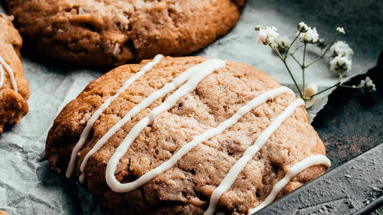 A close-up of a large cookie with white icing drizzled across the top, placed on a textured surface. A small sprig of white flowers rests beside it, reminiscent of Grandma's secret recipes. Other cookies from her cherished fall collection are partially visible in the background.