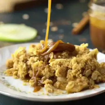 A slice of apple crumble sits on a decorative plate, perfect among Thanksgiving dishes. Caramel sauce is being drizzled over the top, adding just the right touch of sweetness. In the blurred background, a jar of caramel sauce and apple slices await their turn to enhance this delightful treat.