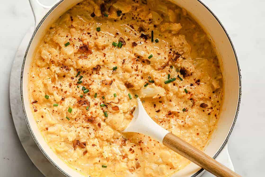 A pot of creamy mashed potatoes garnished with chopped chives and a sprinkle of red pepper flakes. This new favorite is served with a wooden spoon, ready to share.
