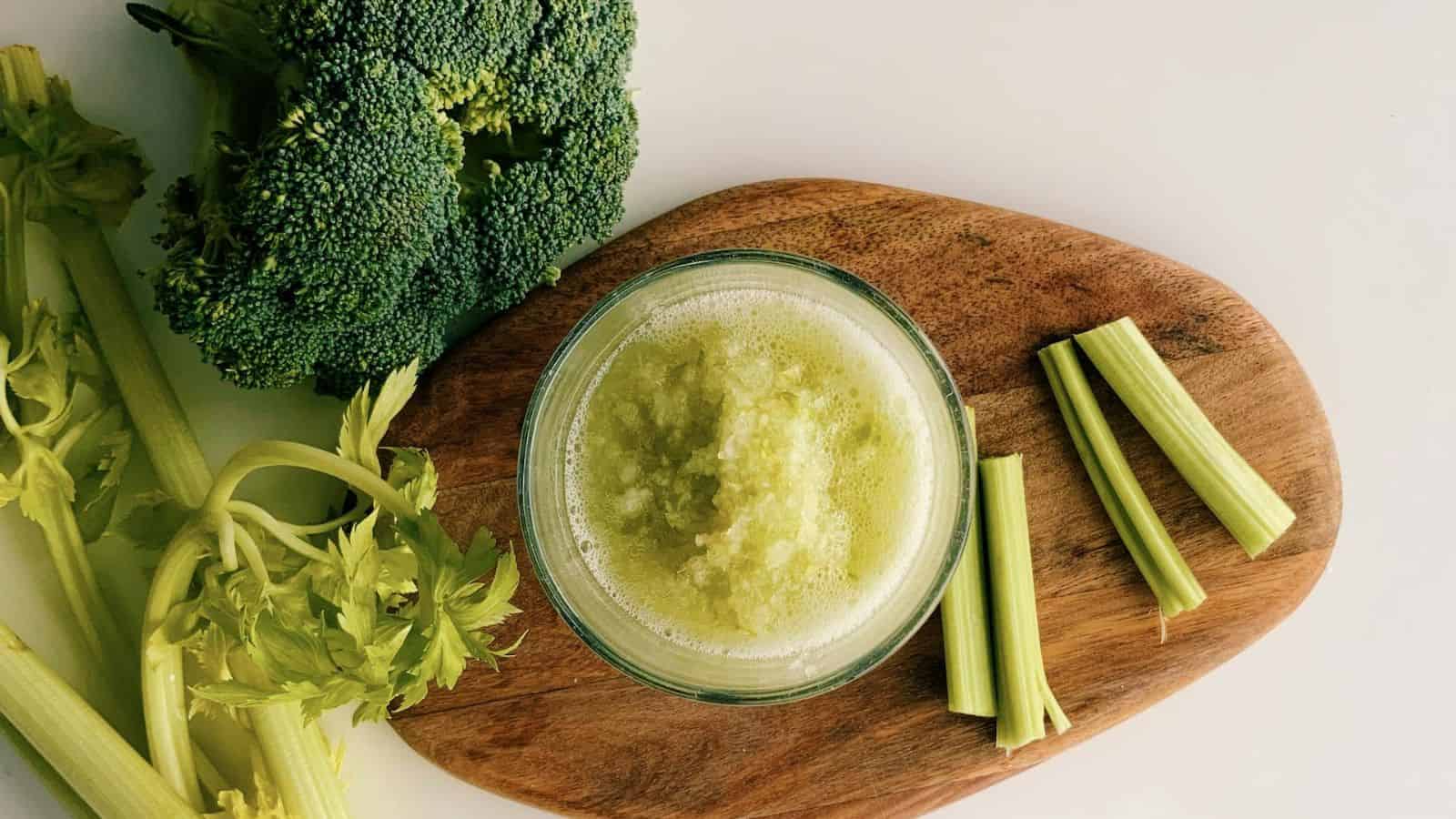 A glass of green smoothie on a wooden board, surrounded by fresh celery stalks and a head of broccoli on a white background.