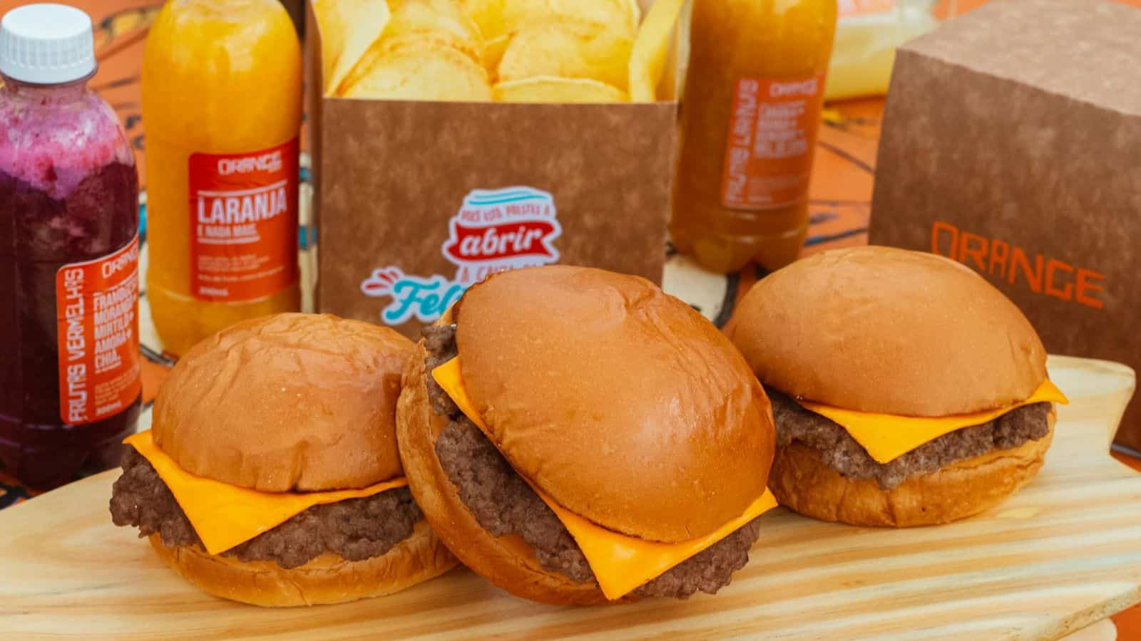 Three cheeseburgers with beef patties and cheddar cheese on buns are displayed on a wooden board. Behind them are a box of potato chips and bottles of juice, labeled "Orange" and "Laranja.
