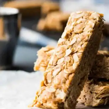 A delectable stack of granola bars with visible oats rests on a crumpled piece of parchment paper. In the blurred background, a metal cup sits beside more bars, offering a glimpse of one of the 15 no-bake desserts perfect for any sweet craving.