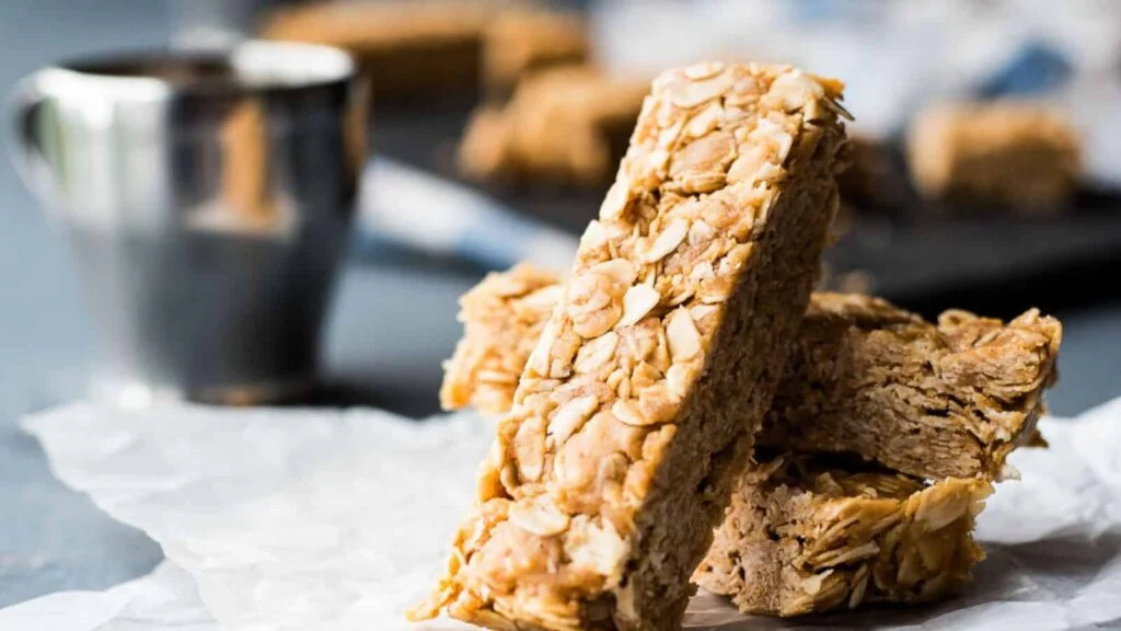 A delectable stack of granola bars with visible oats rests on a crumpled piece of parchment paper. In the blurred background, a metal cup sits beside more bars, offering a glimpse of one of the 15 no-bake desserts perfect for any sweet craving.