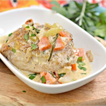 A cooked piece of meat is served on a white rectangular dish, topped with diced vegetables and a creamy sauce. This favorite recipe is placed on a wooden board, with a colorful patterned cloth partially visible in the background, making it an inviting find for any culinary enthusiast.