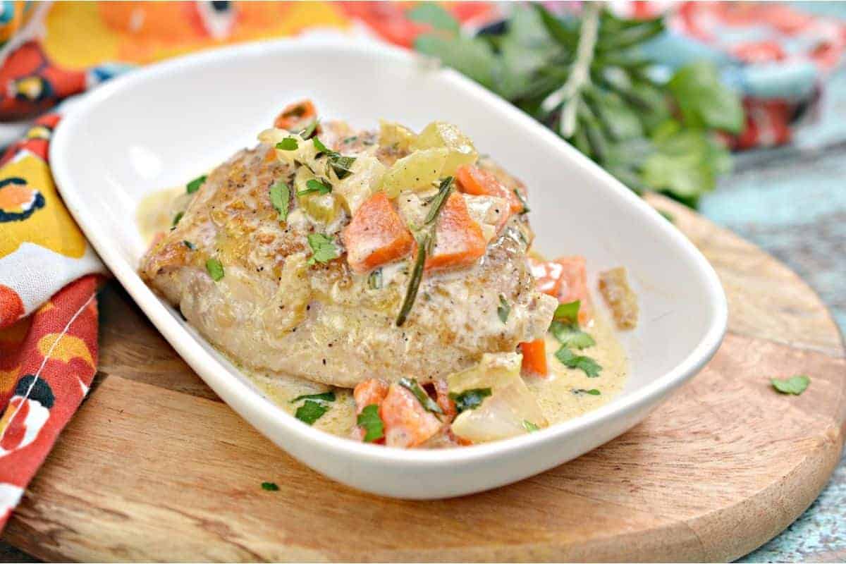 A cooked piece of meat is served on a white rectangular dish, topped with diced vegetables and a creamy sauce. This favorite recipe is placed on a wooden board, with a colorful patterned cloth partially visible in the background, making it an inviting find for any culinary enthusiast.