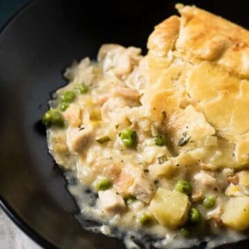 A serving of chicken pot pie is presented in a black bowl, perfect for a Thanksgiving without turkey. The pie boasts a golden, flaky crust with a filling of chicken, peas, and potatoes in creamy sauce. A white napkin peeks from beneath the bowl.