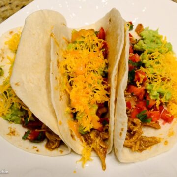 Two soft tacos, the ultimate game day snacks, are filled with shredded chicken, diced tomatoes, shredded cheese, and guacamole placed on a white plate. Tasty as ever!