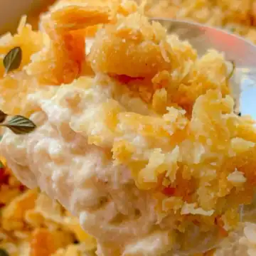 A close-up of a spoonful of creamy, cheesy potato casserole topped with a golden, crispy crust that looks good enough for an Auto Draft. The casserole is garnished with herbs, visible in the background. The texture appears rich and crunchy.