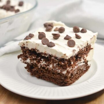 A close-up image of a carb-loving slice of chocolate cake with white frosting and chocolate chips on top, served on a white plate. The cake is layered and placed on a wooden surface with a white napkin in the background.