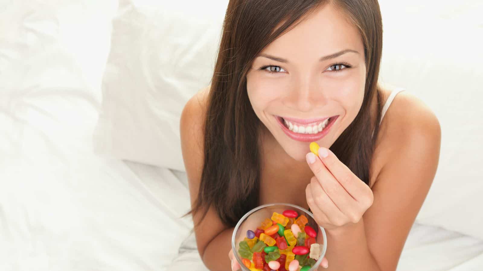 A person with long hair is smiling while holding a bowl of colorful jelly beans. They are sitting on a white surface and holding one yellow jelly bean in their hand.