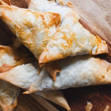 Golden-brown triangular pastries are arranged on a wooden surface. The pastries have a flaky, crisp texture with visible layers of dough. Crumbs and small pieces of pastry are scattered around, offering a perfect example of quick recipes made to impress.