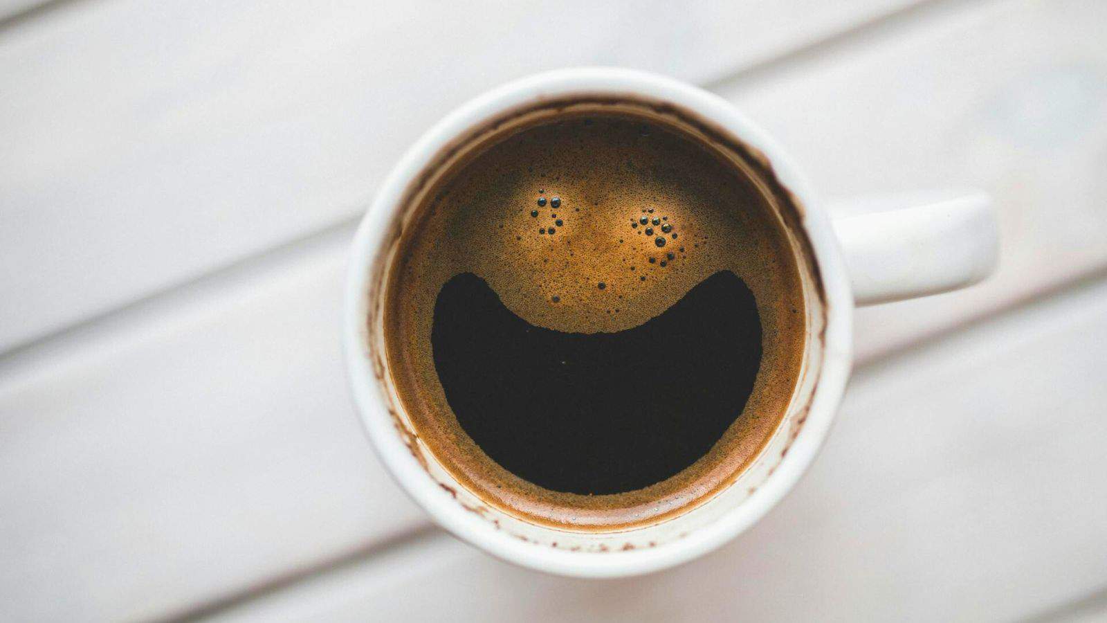 A top-down view of a white cup filled with coffee. The bubbles form a pattern resembling a smiling face. The cup is on a white wooden surface.