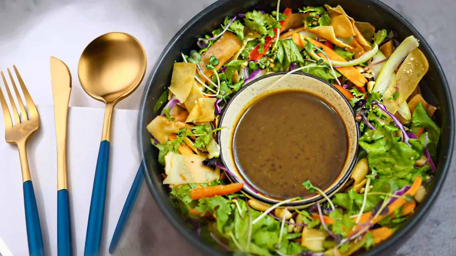 A bowl of fresh salad with mixed greens, shredded carrots, sliced red peppers, and crispy pita chips, centered with a bowl of brown dressing. Next to it, gold cutlery with navy blue handles is placed on a white napkin.