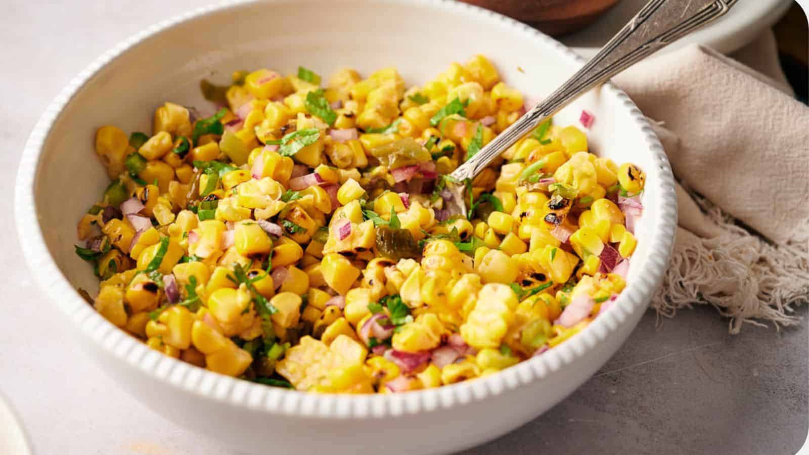 A white bowl brimming with a vibrant corn salad, complete with grilled corn kernels, red onion, green peppers, and herbs&mdash;an ideal addition to your recipes for Thanksgiving sides. A spoon rests in the bowl against a backdrop of a soft fabric napkin. Perfect for impressing your in-laws!