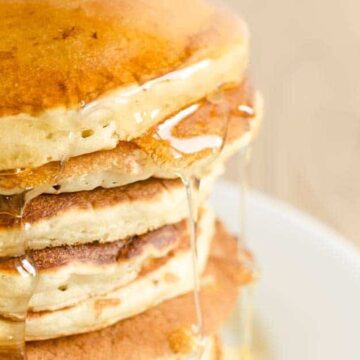 A stack of five fluffy pancakes is topped with syrup, cascading down the sides like an auto draft of sweetness. The pancakes rest on a pristine white plate.