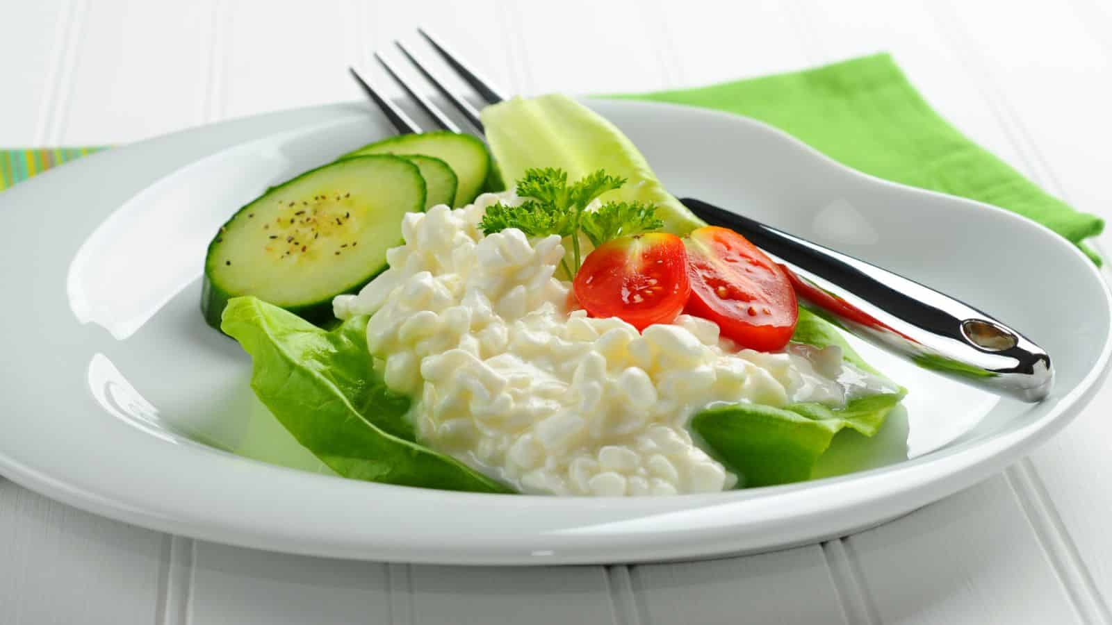 A plate with cottage cheese on a bed of lettuce, garnished with parsley. Sliced tomatoes and cucumber are on the side. A fork and knife are placed next to the food on a white plate. A green napkin is beneath the plate.