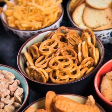 A curious assortment of international snacks, featuring pretzels, breadsticks, peanuts, cracker sticks, and chips, fills various bowls arranged on a dark surface. This weird yet delightful spread offers a taste adventure for all snack enthusiasts.
