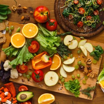 A wooden table is artfully arranged with fresh fruits and vegetables, reminiscent of an Auto Draft masterpiece. A large cutting board showcases sliced apples, oranges, tomatoes, peppers, lettuce, and herbs. Plates nearby brim with salads and diced produce, accented by scattered walnuts and grapes.