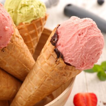 Four waffle cones filled with ice cream are arranged in a wooden bowl. The ice cream flavors include pink and light green. A scoop of pink ice cream, berries, and mint leaves are visible on a wooden surface in the background.