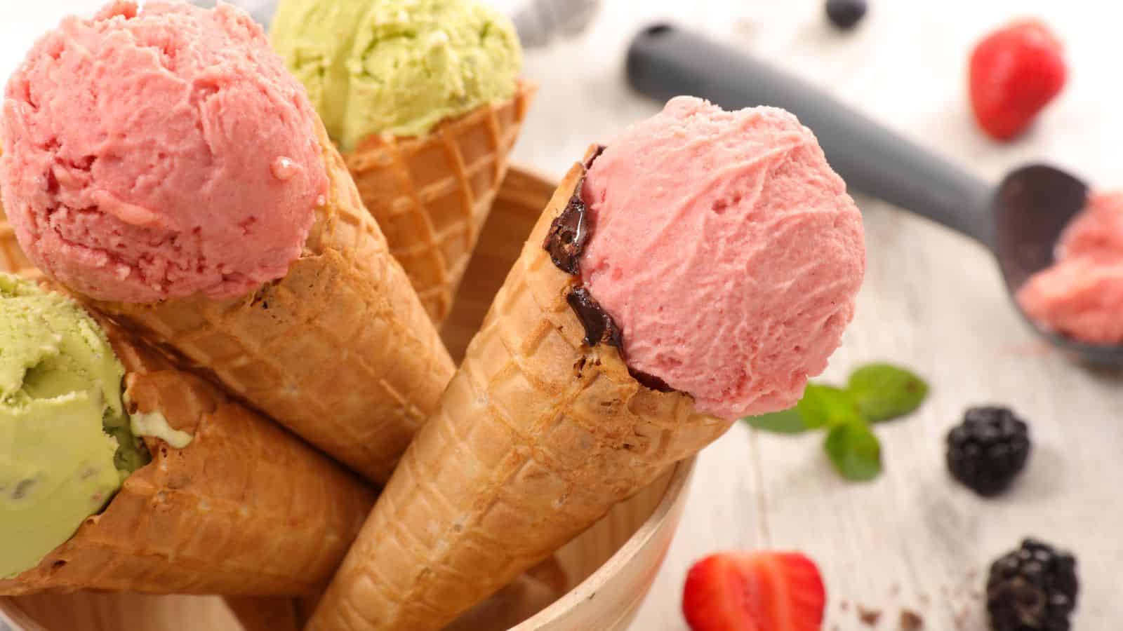 Four waffle cones filled with ice cream are arranged in a wooden bowl. The ice cream flavors include pink and light green. A scoop of pink ice cream, berries, and mint leaves are visible on a wooden surface in the background.