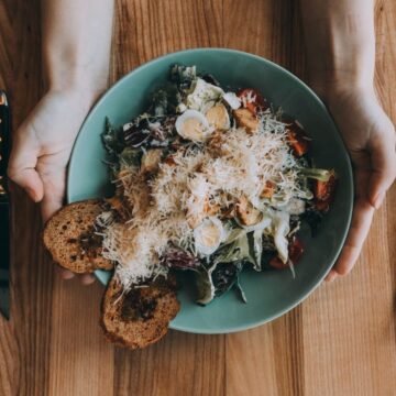 A person cleverly makes the most of leftover ingredients, holding a green plate with a salad topped with shredded cheese and two slices of brown bread. A knife rests on the left and a fork sits on the right, all arranged neatly on a wooden table.