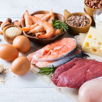 A variety of protein-rich foods arranged on a white wooden surface, including raw salmon, shrimp, beef, chicken breast, eggs, cheese, nuts, seeds, grains, cottage cheese, and a small pitcher of milk.