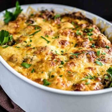 A baked casserole in a white oval dish, topped with melted cheese and garnished with parsley, offers a delightful Thanksgiving without turkey option. It sits on a brown cloth with a serving spoon and fork nearby.