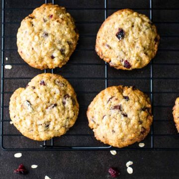 Six of Grandma's secret oatmeal cookies with cranberries and chocolate chips cool on a black wire rack atop a dark surface. Scattered oats and cranberries add to the charm, making it a timeless favorite among fall cookie recipes.
