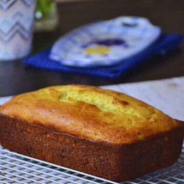 A loaf of banana bread sits on a cooling rack. In the background, there is a patterned mug and a decorative plate on a blue napkin. The setting is on a wooden table with soft lighting.