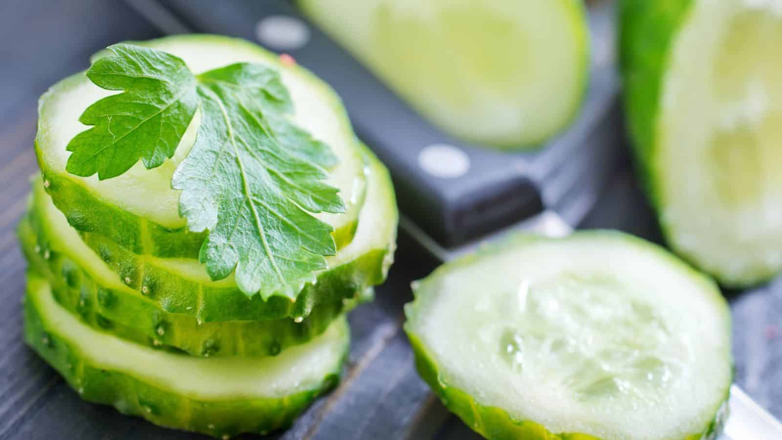 Close-up of sliced cucumbers arranged on a dark surface. A parsley leaf is placed on top of the stack of cucumber slices. A blurred knife is visible in the background, along with more cucumber slices.