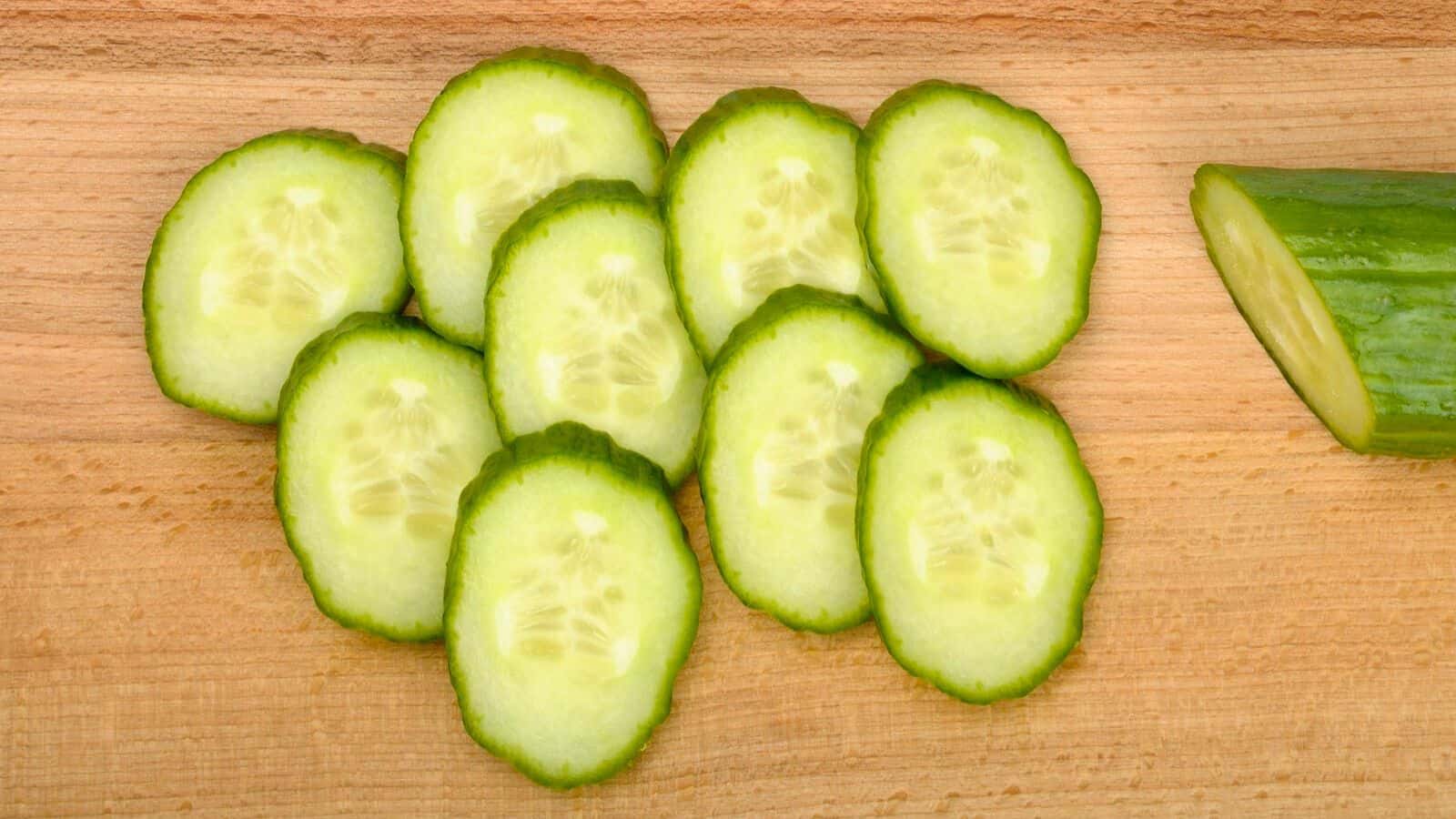 Slices of cucumber are neatly arranged in an overlapping pattern on a wooden cutting board. A partial view of an unsliced cucumber is visible on the right side of the board.