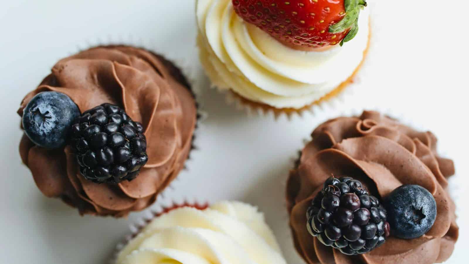 Four cupcakes with swirled frosting are arranged. Two have chocolate frosting topped with a blackberry and a blueberry. One has vanilla frosting with a strawberry, and one has vanilla frosting without fruit. They sit on a white surface.