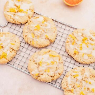 Six glazed cookies with orange zest are arranged on a cooling rack. Slices of orange are placed around the cookies on a light-colored surface.