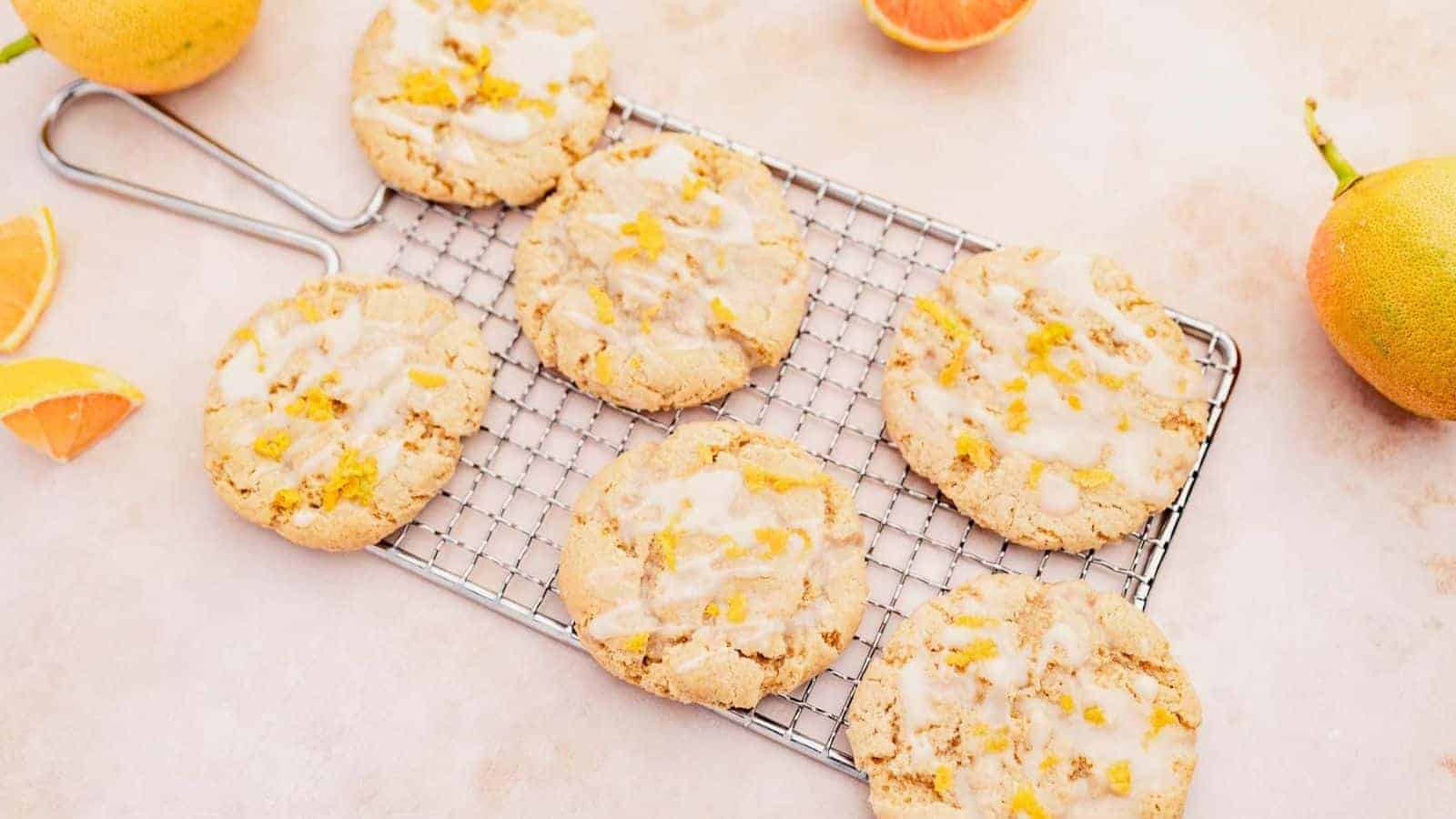 Six glazed cookies with orange zest are arranged on a cooling rack. Slices of orange are placed around the cookies on a light-colored surface.