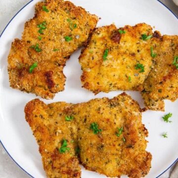 A white plate with four pieces of breaded and fried chicken, garnished with parsley. A beige cloth is on the right, and a sprig of fresh parsley is partially visible in the top left corner.